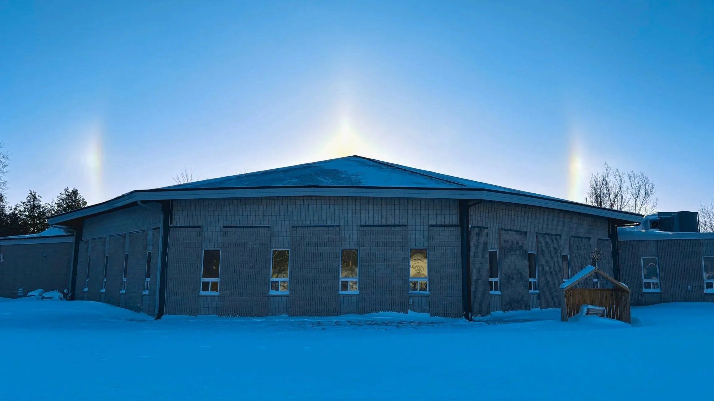 Broadway Pentecostal Church in winter, a long single-storey building of grey block under a broad hipped roof, with a row of windows above the snow and sun dogs in a clear sky