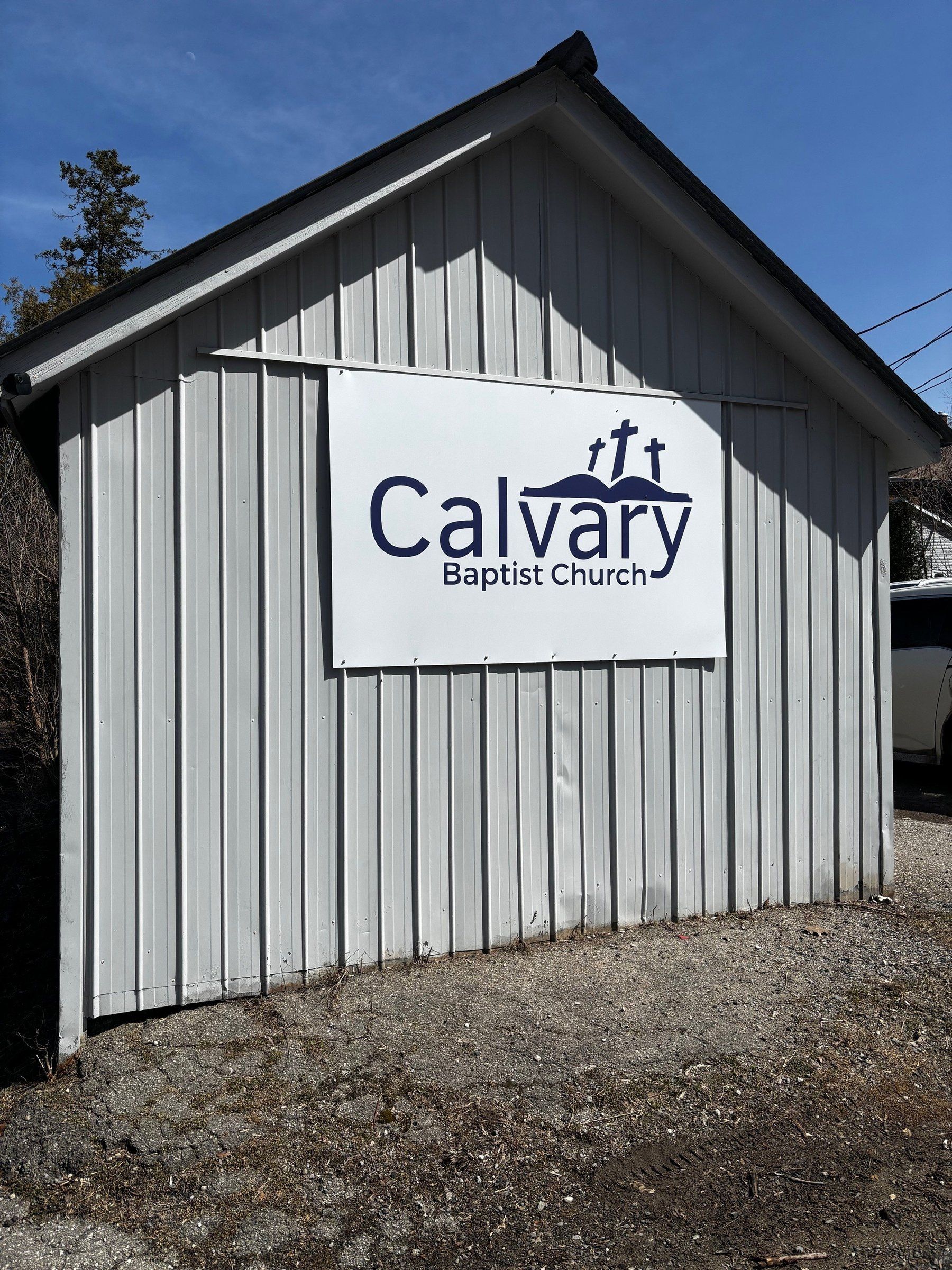 The gable end of Calvary Baptist Church's hall on Penny Lane, clad in pale grey vertical steel siding, with a large white sign bearing the church's name and three crosses