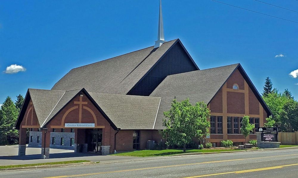 Orangeville Canadian Reformed Church, a brick church with a tall shingled roof and a slender white spire, and a covered entrance under a gable marked with a cross
