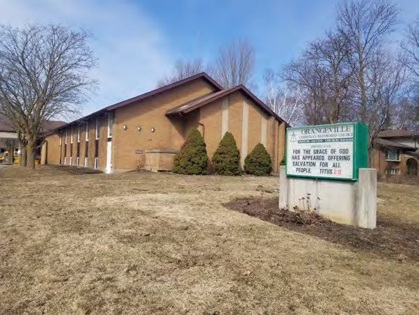 Orangeville Christian Reformed Church, a long low buff-brick church with a row of tall narrow windows and a gabled front, behind a green roadside sign bearing its name, in early spring