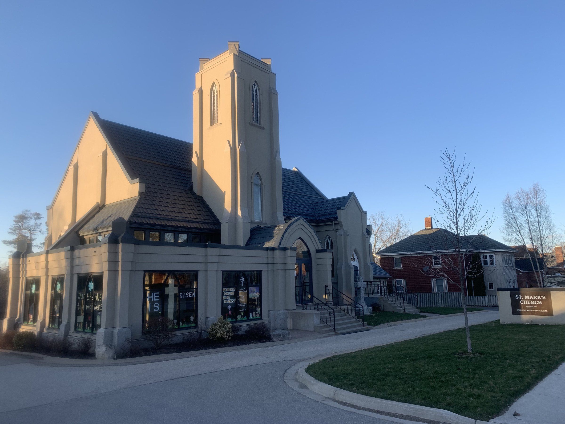 St. Mark's Anglican Church in evening light, a cream-rendered Gothic church with a square tower, a dark tiled roof and a one-storey glazed corner wing, beside its roadside sign