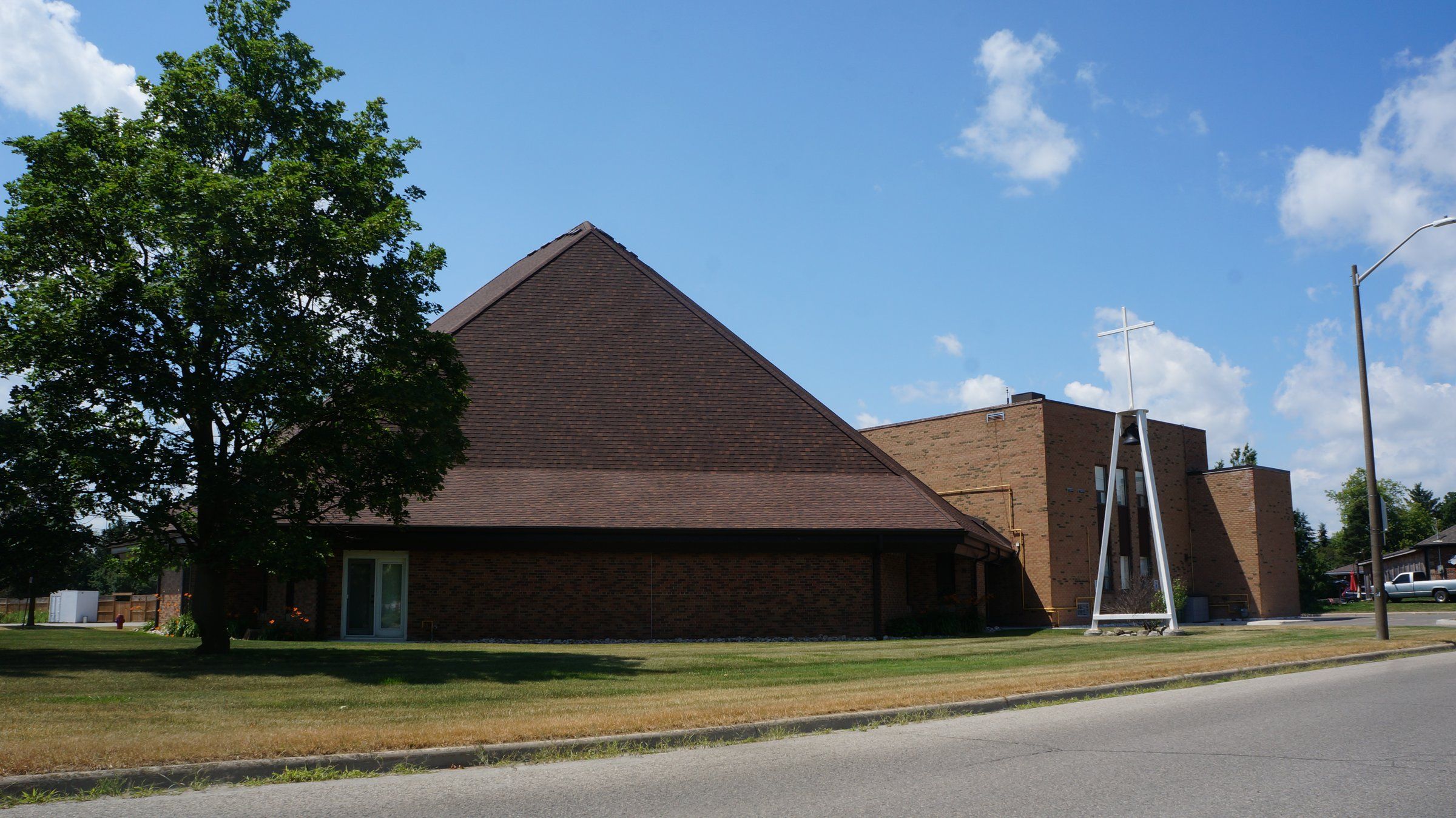 St. Timothy Catholic Church, a brick church under a broad, steep shingled roof, with a taller brick block behind it and a white steel frame holding a bell beneath a cross