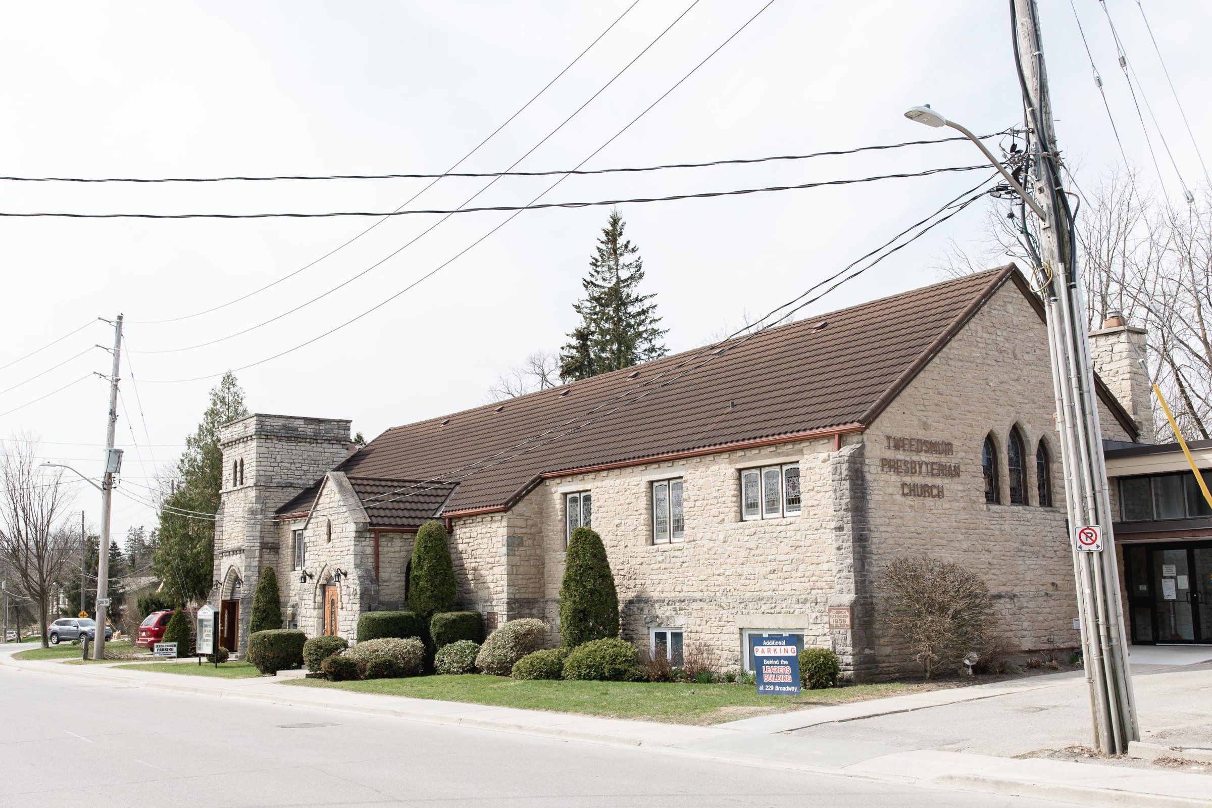 Tweedsmuir Memorial Presbyterian Church, a long buff stone church with a brown tile roof and a square crenellated tower over an arched entrance, its name lettered on the end gable, seen from John Street
