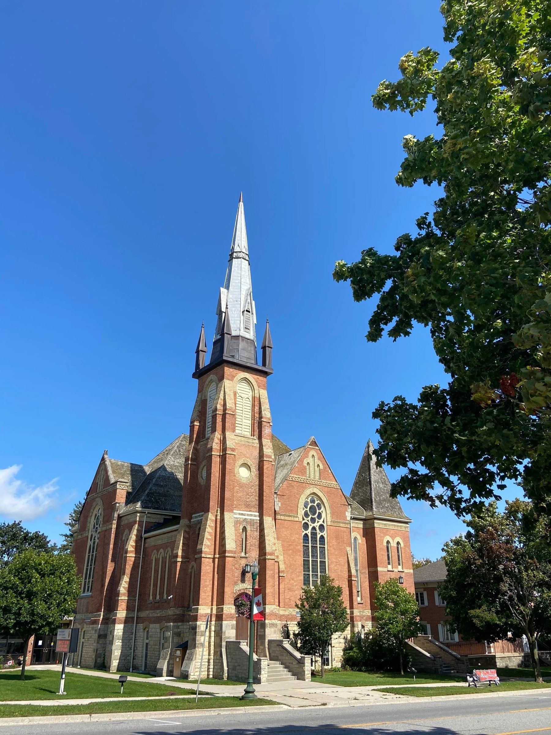 Westminster United Church, a red-brick Gothic church with buff-brick trim and a rose window, its central tower topped by a tall metal-clad spire ringed with four pinnacles, seen from Broadway on a sunny day